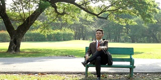 亚洲年轻的商人坐在一个公园的木凳上放松