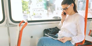 一名年轻女子在有轨电车里一边用平板电脑一边用手机说话。