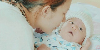 快乐的亚洲母亲抱着和亲吻她的混血可爱的小新生儿的脸颊在她的卧室