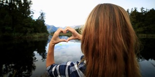 一个年轻女子站在湖边，在风景上做了一个心形的手指框架