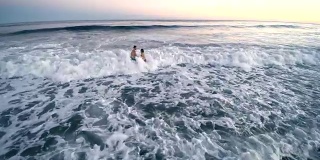 日落时分，一名年轻男子和一名少女在海里游泳，被海浪击中。汉普顿海滩，长岛，纽约州，美国。空中低空视频。