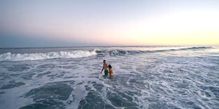 日落时分，一名年轻男子和一名少女在海里游泳，被海浪击中。汉普顿海滩，长岛，纽约州，美国。