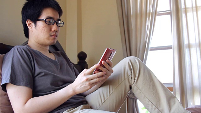 一名亚洲男子戴着眼镜在床上用手机发短信