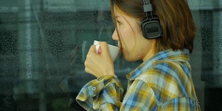 女士喝咖啡和听音乐。外面在下雨，侧视图。从右到左。