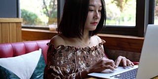 年轻微笑的亚洲女孩正在玩笔记本电脑和享受咖啡在咖啡馆