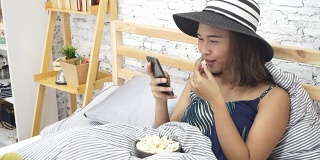 Beautiful young Asian women touching smart phone tablet on bed and eating pop corn