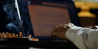 一个男人在笔记本电脑上工作，在一个装满烟头的烟灰缸上抽烟