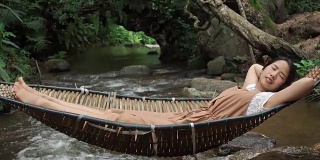 亚洲女人放松睡在吊床上的自然森林与小溪