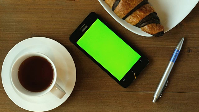 早间新闻与咖啡-咖啡杯和巧克力羊角面包在木桌子上绿色屏幕智能手机。