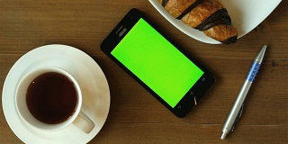 早间新闻与咖啡-咖啡杯和巧克力羊角面包在木桌子上绿色屏幕智能手机。