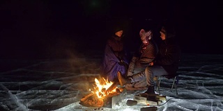 三个旅行者在夜间的冰面上着火。营地在冰上。帐篷紧挨着火。贝加尔湖。附近有一辆车。人们围着篝火取暖，穿着睡袋。