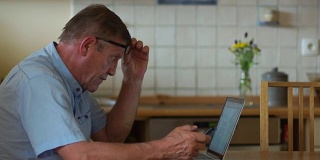 为退休人员提供的互联网技术。一位上了年纪的人正坐在餐桌前用笔记本电脑工作。他举起眼镜，向屏幕倾斜，这样能更好地看数字。近视