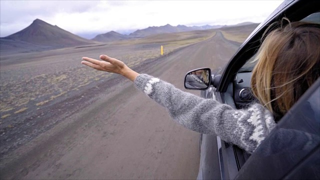 开朗的白种女性在车里看着路伸出双臂，山上火山景观。公路旅行的概念。金发女孩看着风景。慢动作