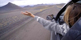 开朗的白种女性在车里看着路伸出双臂，山上火山景观。公路旅行的概念。金发女孩看着风景。慢动作