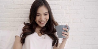美丽的亚洲女人玩电脑或笔记本电脑，而躺在床上在她的卧室。快乐的女性喝咖啡，同时在家里网购。生活方式女性在家的概念。