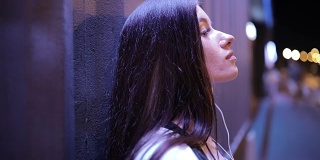 一个女人戴着耳机听着音乐，靠在夜城的墙上放松