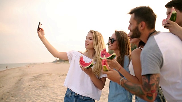 朋友们站在沙滩上吃西瓜，用智能手机相机自拍。年轻的男人和女人穿着蓝色的牛仔裤短裤在海边聊天。健康营养，素食