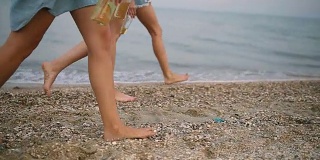 孤立的观点，三对苗条的女性腿与玻璃瓶的饮料在手踩在海边的海滩在日落慢动作。年轻的女人在沙滩上喝着啤酒。没有脸