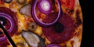 开胃的自制披萨，有意大利腊肠、泡菜、橄榄、西红柿、奶酪和洋葱圈，刚从烤箱中取出，切下来的一块在黑色背景上旋转