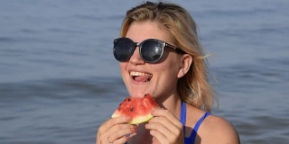 一个戴墨镜的女人坐在海滩上吃西瓜。一个金发女郎手里拿着一个成熟的西瓜