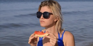 一个戴墨镜的女人坐在海滩上吃西瓜。一个金发女郎手里拿着一个成熟的西瓜