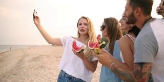 朋友们站在沙滩上吃西瓜，用智能手机相机自拍。年轻的男人和女人穿着蓝色的牛仔裤短裤在海边聊天。健康营养，素食
