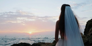 不认识的女孩穿着豪华的婚纱在海边看日出。石头上的新娘。美丽的波浪靠近她。女人享受与自然的快乐时光。慢动作