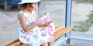 一个带着帽子和背包的小女孩正坐在公交车站的长椅上，等待进一步旅行的交通工具