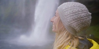 女游客在冰岛旅游的慢动作视频站在壮观的瀑布旁边，沉思着大自然，人们旅行冒险的概念