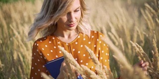 美丽快乐的年轻女子在田间的小穗和小麦上带着书的夕阳，金发碧眼的在草地上度过了美好的时光