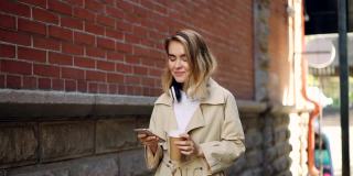 慢镜头中，一名快乐的年轻女子用智能手机走在街上，带着外带咖啡和耳机。现代科技，饮料和人的概念。