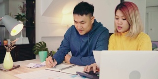 在现代家庭中，年轻的亚洲夫妇使用笔记本电脑和计算器管理财务，查看他们的银行账户。女人和男人一起做文书工作，用笔记本电脑在线纳税。