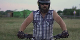 英俊的男子摩托车手与他的头盔和定制摩托车在野外日落