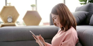 一名亚洲妇女躺在客厅的沙发上使用平板电脑。快乐的女性在家用平板电脑发短信、阅读、看视频和上网。生活方式女性在家的概念。