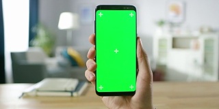 一个男人的特写持有绿色模拟屏幕智能手机和使用触摸屏手势。触摸手机色度键屏幕。在背景舒适的家庭气氛。