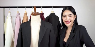 快乐微笑的亚洲女性裁缝时装设计师与人体模型假人和西装夹克在许多颜色的衣架