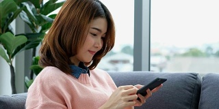 年轻美丽的亚洲女人使用智能手机与幸福，坐在沙发上与城市的看法，人们在电话，生活方式