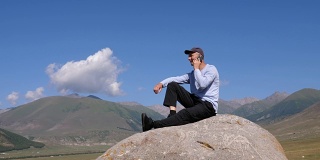 男人坐在山上的大石头上用手机打电话