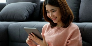 年轻美丽的亚洲女人使用数字平板电脑与幸福坐在沙发上与城市的看法，人们在电话，生活方式