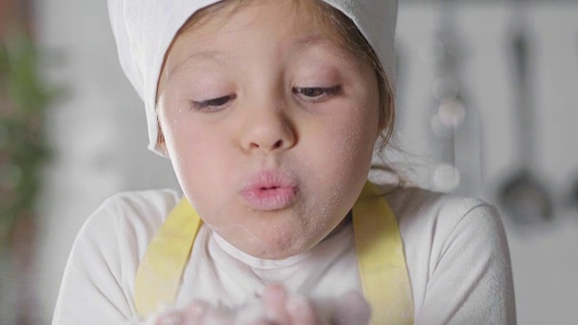 一个小女孩打扮成专业厨师在厨房里玩面粉，弄脏了她的手，开心地笑了。