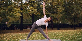 有魅力的年轻女子正在户外做瑜伽，站在垫子上，练习一系列体式，三角姿势和勇士姿势，享受新鲜的空气。