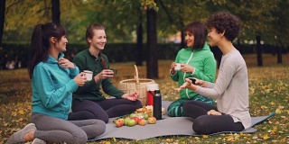 快乐的女孩们在公园里野餐，坐在瑜伽垫上，在秋天的户外练习后吃东西，女孩们有说有笑。沟通与美食理念。