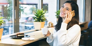 一个美丽快乐的女人，坐在靠窗的咖啡馆里，一边拿着一杯咖啡一边打电话