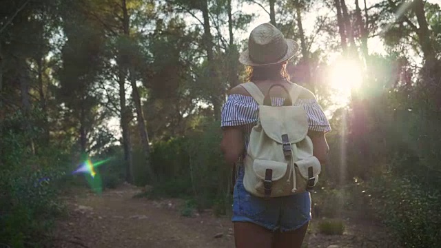在一个阳光明媚的日子里，一幅美丽的年轻游客(女孩)的肖像，背着背包，戴着草帽，走过森林，背景是树木。