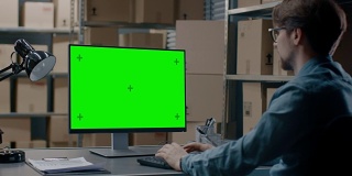 仓库库存经理坐在办公桌前，在一台绿色模拟屏幕电脑上工作。在背景货架上装满了纸箱包装准备运输。