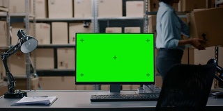 在仓库计算机与绿色模拟屏幕显示站在桌面上。在背景的货架上装满了纸箱和包裹的产品准备运输。