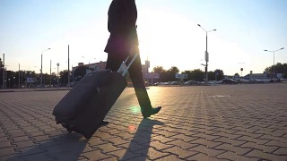 简介成功的商人在一个黑色西装走在他的行李在城市街道日落。一个自信的年轻人拖着带轮子的行李箱走向机场。关闭侧视图视频素材模板下载