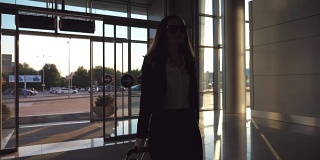 商务女士带着行李去机场。穿着高跟鞋的年轻女子穿过玻璃门进入航站楼，用轮子推着行李箱。旅行或度假旅游概念。慢镜头