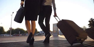 商务人士离开机场，带着行李走在城市街道上。穿着高跟鞋的陌生男人和女人从航站楼走出来，用轮子滚动行李箱。出差的概念。慢镜头后视图特写
