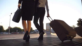 商务人士离开机场，带着行李走在城市街道上。穿着高跟鞋的陌生男人和女人从航站楼走出来，用轮子滚动行李箱。出差的概念。慢镜头后视图特写视频素材模板下载